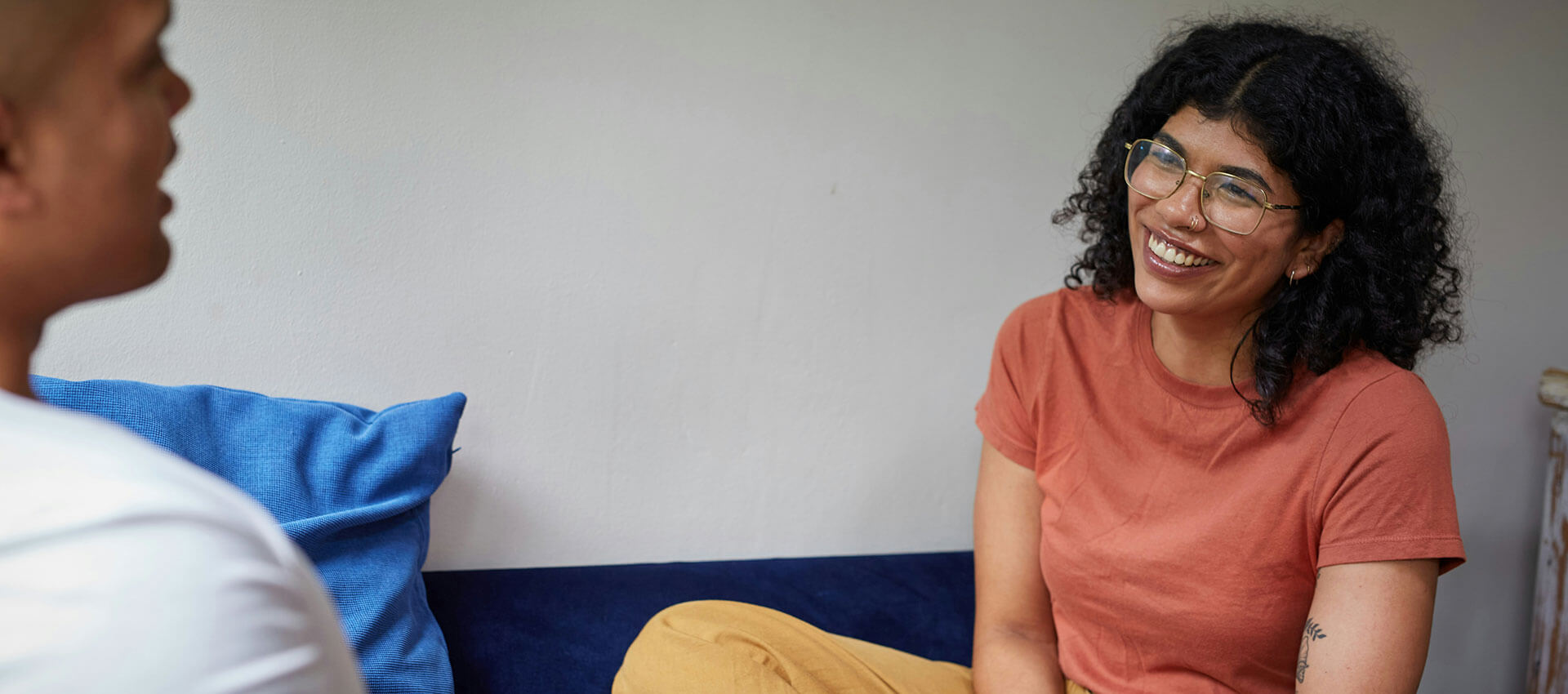A woman with curly hair and glasses smiles while talking to a person off-camera, sitting in a room with a blue cushion and neutral-toned walls.