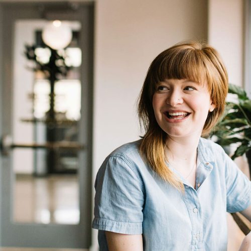 A person is sitting and smiling by a window with natural light, next to a potted plant on a wooden surface in a modern indoor setting.