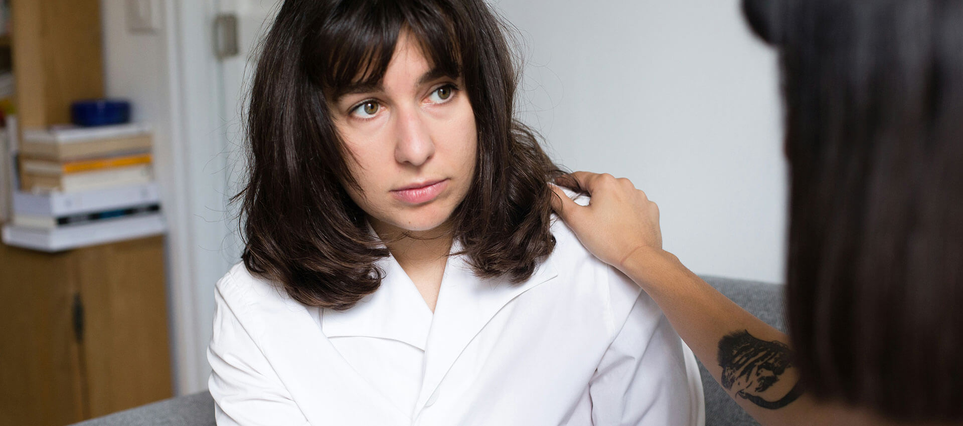 A person in a white shirt sits on a couch, receiving a comforting hand on their shoulder from another person with a tattooed arm, in a cozy room with shelves.