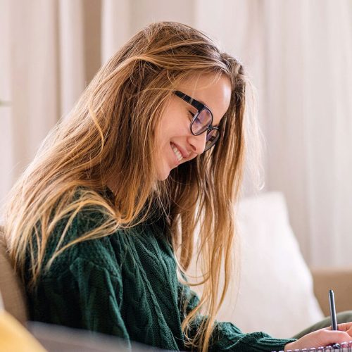 A woman with long hair and glasses sits on a couch, writing in a notebook. She is in a bright, cozy room with a potted plant in the background.