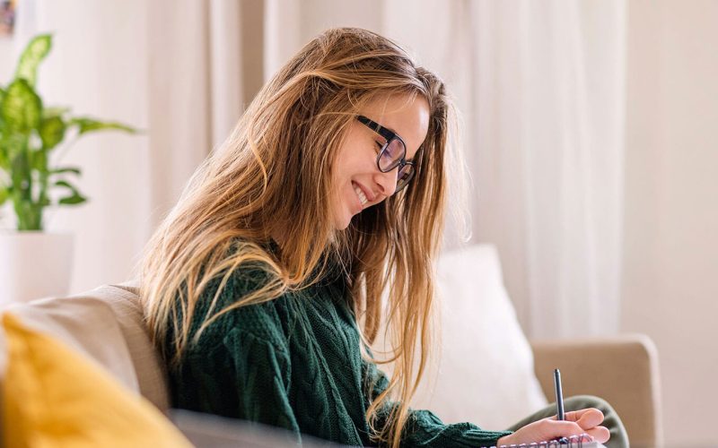 A woman with long hair and glasses sits on a couch, writing in a notebook. She is in a bright, cozy room with a potted plant in the background.