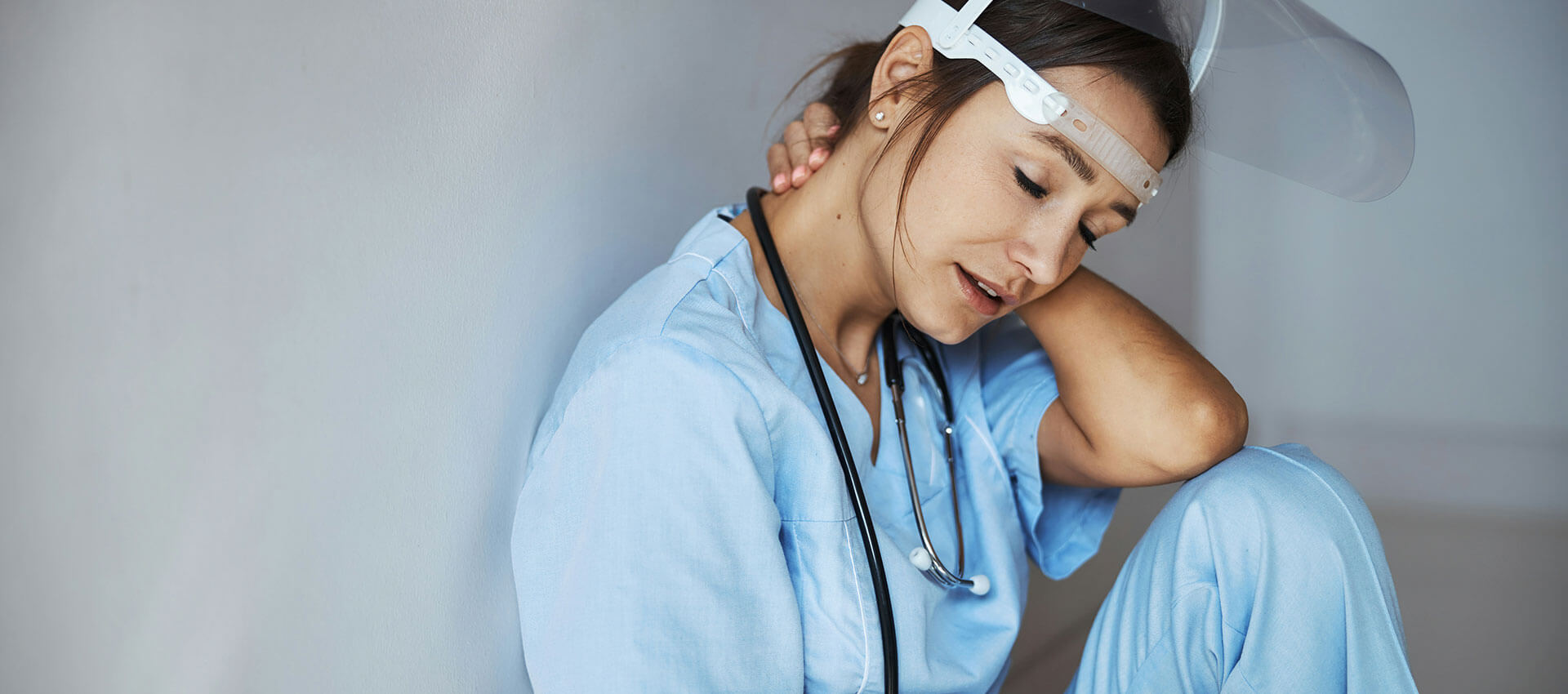 A healthcare worker in blue scrubs, wearing a face shield and stethoscope, sits against a wall, massaging her neck with a tired expression in a clinical setting.