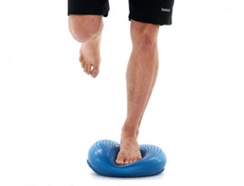 Person balances on one leg atop a blue inflatable cushion, demonstrating stability exercises. The individual wears black shorts, with the background being plain white.