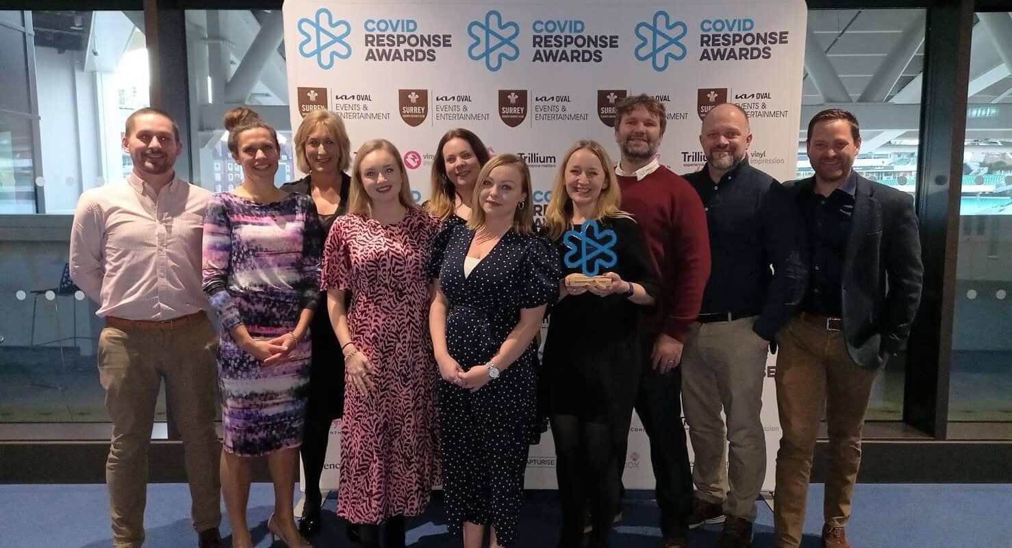 A group of ten people stands smiling in an indoor setting. One person holds an award. Behind them is a backdrop with logos, including "COVID Response Awards" and "Kia Oval Events & Entertainment."