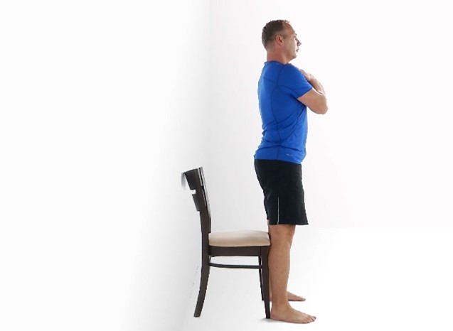 Man in blue shirt and black shorts stands with back against a wall, arms crossed, in front of a chair in a white room.