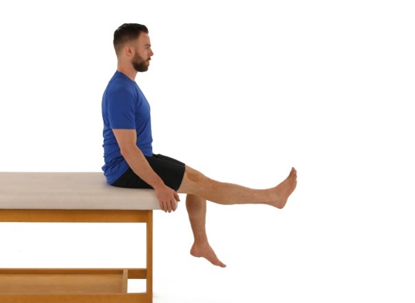A man in a blue shirt and black shorts sits on a therapy table, raising one leg forward in a neutral, white background.