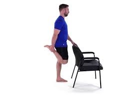 Man stretches quadriceps by holding one foot behind, balancing with free hand on a chair. He is wearing a blue shirt and black shorts, standing on a plain white background.