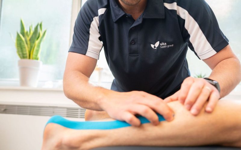 Person in Vita Health Group polo shirt applies blue kinesiology tape to a patient's leg in a well-lit room with potted plants on the windowsill.