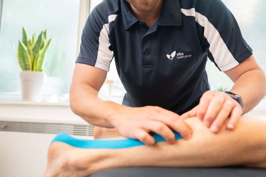 Person in Vita Health Group polo shirt applies blue kinesiology tape to a patient's leg in a well-lit room with potted plants on the windowsill.