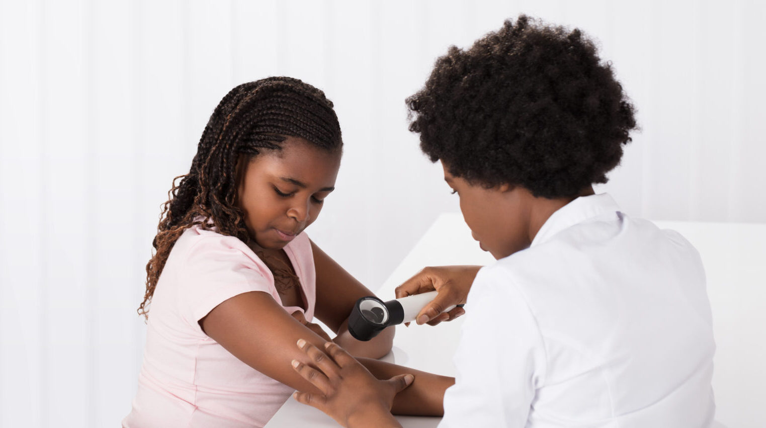 Healthcare professional examines a young patient's arm with a dermatoscope in a clinical setting with a white background.