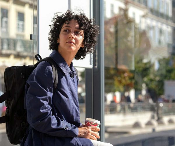 A person with curly hair, wearing a blue jacket and backpack, sits on a bench holding a coffee cup. They look thoughtfully into the distance in an urban setting.