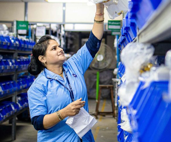 A woman in a blue uniform reaches for items on a high shelf, surrounded by organized rows of blue bins in a warehouse setting.