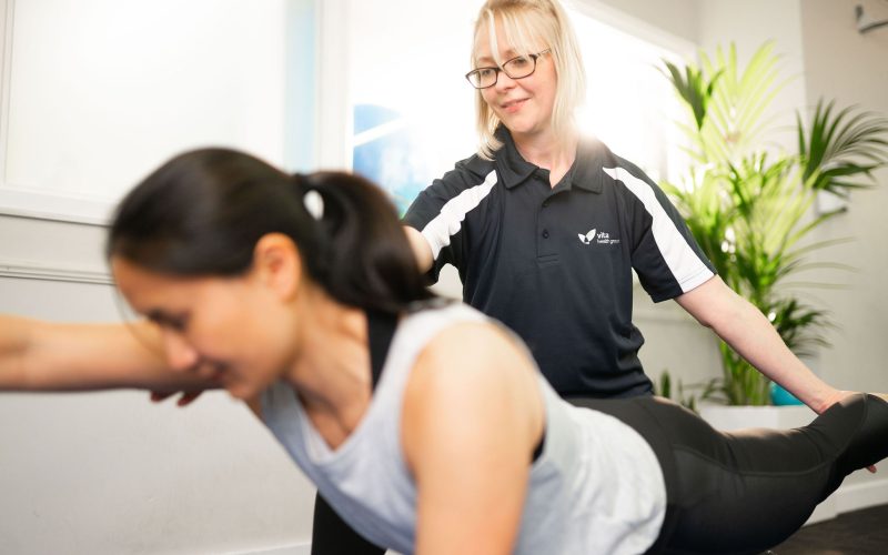 A healthcare professional assists a woman performing an exercise in a bright room with plants. The professional wears a shirt with the logo "Vita Health Group."