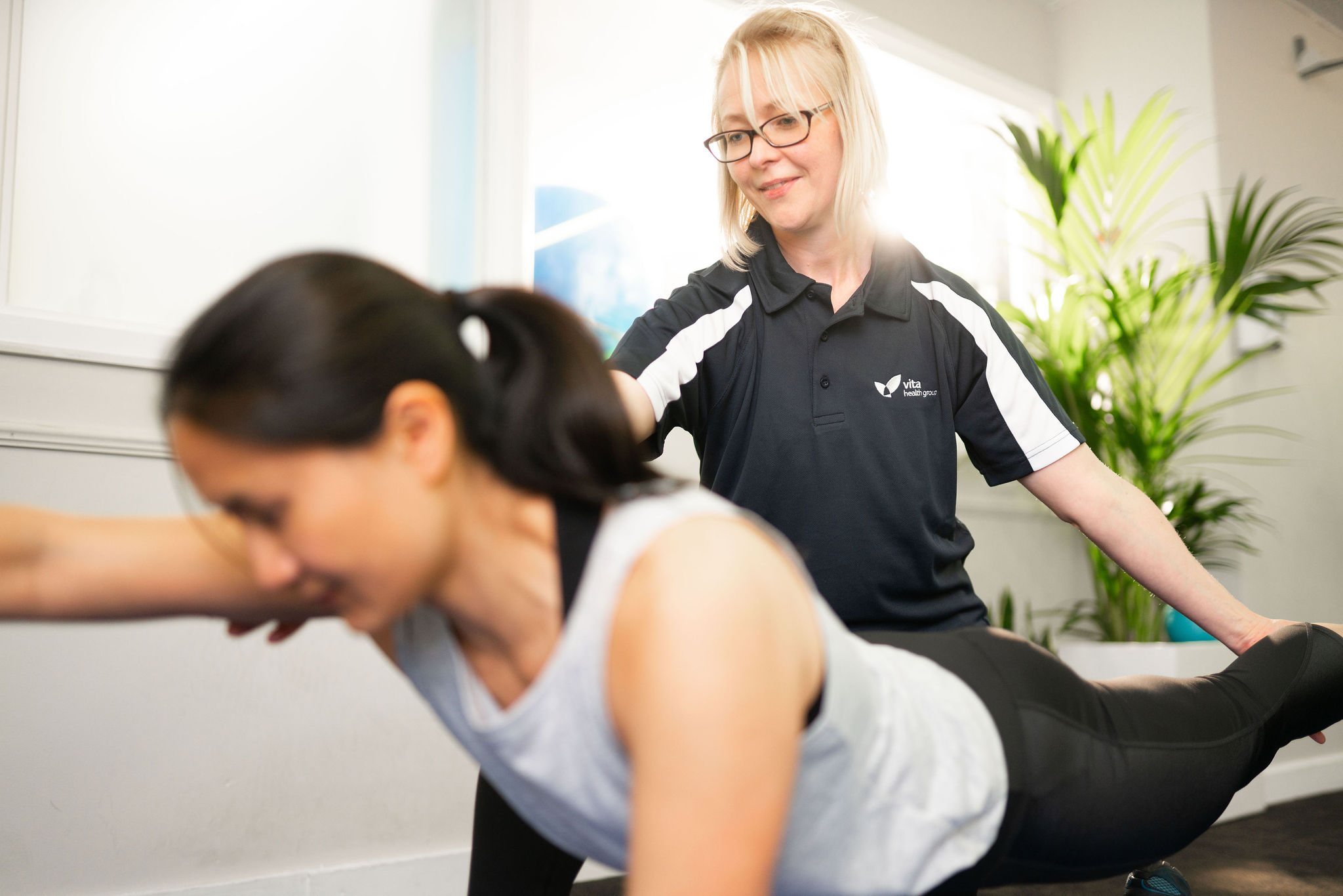 A healthcare professional assists a woman performing an exercise in a bright room with plants. The professional wears a shirt with the logo "Vita Health Group."