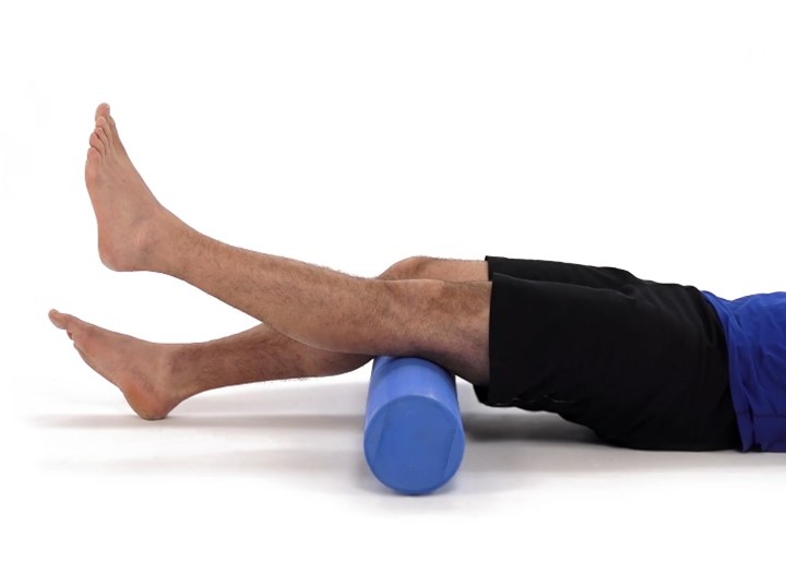 A person is lying on their back using a blue foam roller under their calves. They are wearing black shorts and a blue shirt against a plain white background.