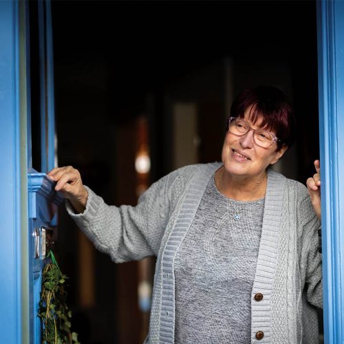 An older woman wearing glasses and a gray cardigan stands smiling in a blue doorway, holding a door handle. The interior appears dimly lit with some greenery on the doorframe.