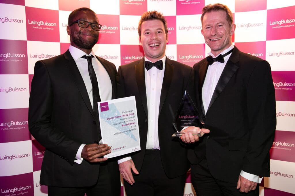 Three men in formal suits stand together smiling. The man on the left holds a certificate reading: 