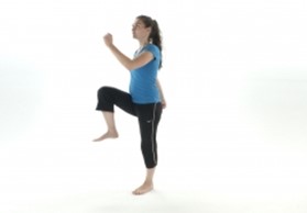 A woman in a blue shirt and black pants performs a high knee exercise, raising one knee while lifting her opposite arm, against a plain white background.