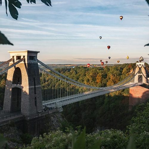 A suspension bridge spans across a deep green valley with several colorful hot air balloons floating in the sky. Trees and foliage surround the bridge under a clear blue sky.