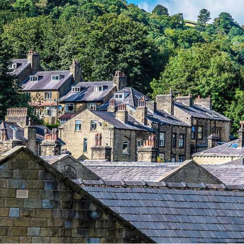 Stone houses with grey slate roofs rest on a hillside, surrounded by lush green trees and a forested background. Sunlight highlights the textures and patterns of the traditional architecture.