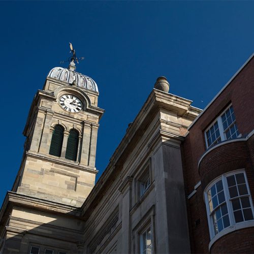 A clock tower with a domed top and weather vane stands prominently against a clear blue sky, adjacent to a red-brick building with rounded windows.