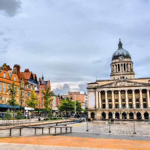 A historic building with a domed clock tower stands adjacent to a row of colorful brick buildings, set around an open, paved city square under a cloudy sky.