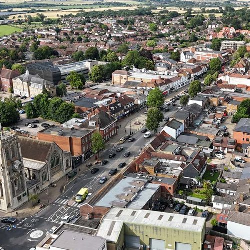 Aerial view of a bustling town with scattered cars on streets. Buildings, including a prominent church, are surrounded by trees, with fields visible in the background.