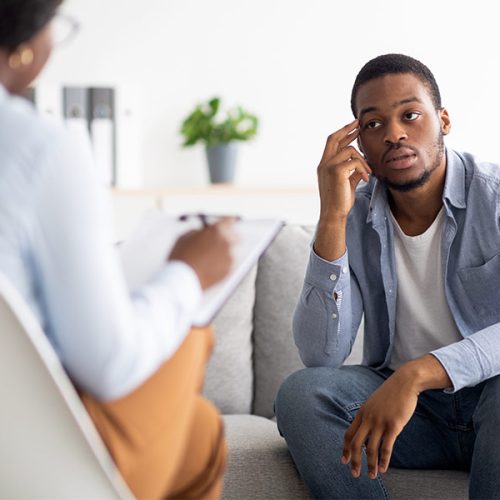 A man sits on a couch, attentively engaging in a conversation with a person taking notes, in a well-lit room with a plant and books in the background.