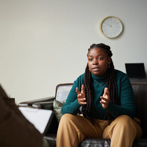 A person in casual clothing sits on a couch, gesturing thoughtfully, while another person takes notes on a clipboard. The room has a potted plant and shelves in the background.