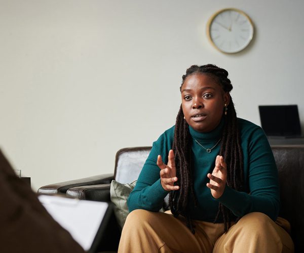 A person in casual clothing sits on a couch, gesturing thoughtfully, while another person takes notes on a clipboard. The room has a potted plant and shelves in the background.