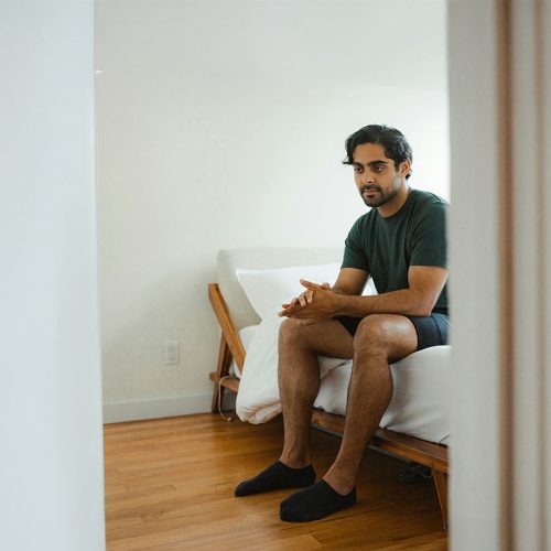A man in a green shirt sits pensively on a bed in a sparsely furnished, bright room with wooden flooring and white walls.