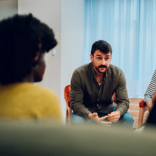 A man in a casual shirt speaks attentively to a group, seated in a room with blue curtains. Two people are partially visible, listening in the foreground.