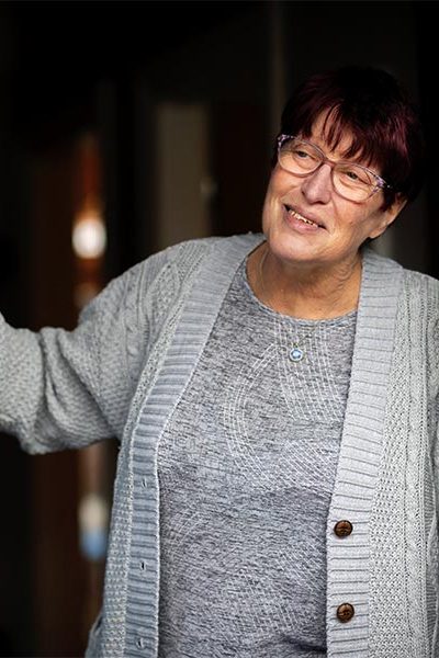 A smiling woman with short hair and glasses is standing indoors, wearing a gray knitted cardigan. She appears relaxed and engaged, with a blurred interior background.