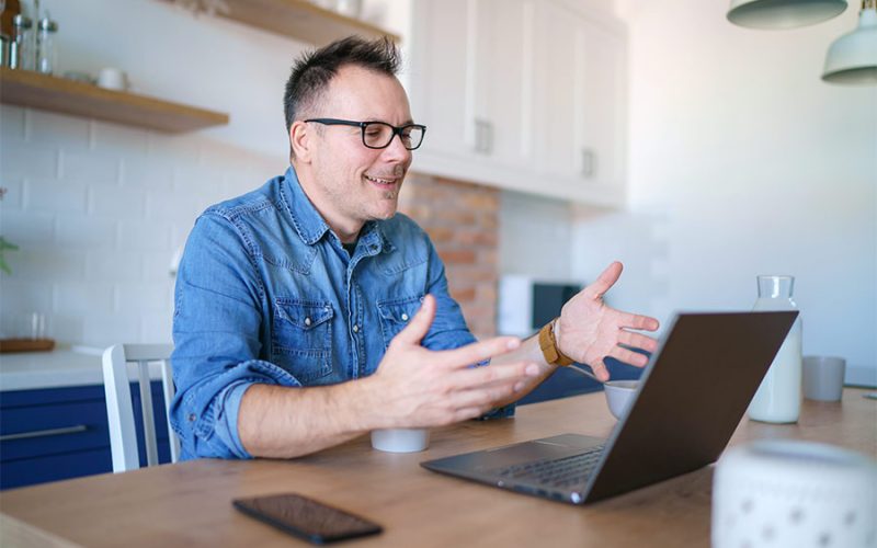 A man in glasses sits thoughtfully with hands clasped, while a woman beside him offers support, placing a hand on his shoulder in a bright room.