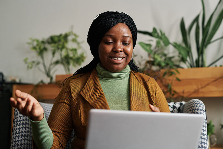 A person is smiling while holding a pen and looking at a laptop screen. They are sitting indoors with green plants and wooden shelves visible in the background.