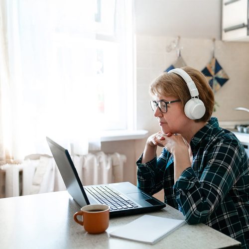 A person wearing headphones and a plaid shirt sits at a table, focused on a laptop. A notebook and mug are nearby in a bright kitchen.