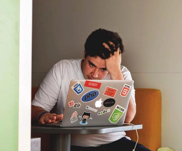 Person seated at a small table, concentrating on a laptop covered in tech-related stickers like GitHub, HTML5, and Node.js, in a modern, casual indoor setting.