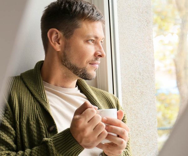 A person holding a white mug gazes thoughtfully out a window, wearing a green sweater. Sunlight illuminates the scene, suggesting a calm, reflective moment in a cozy environment.