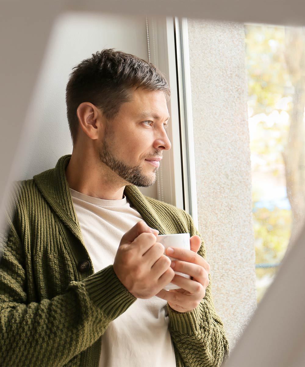 A person holding a white mug gazes thoughtfully out a window, wearing a green sweater. Sunlight illuminates the scene, suggesting a calm, reflective moment in a cozy environment.