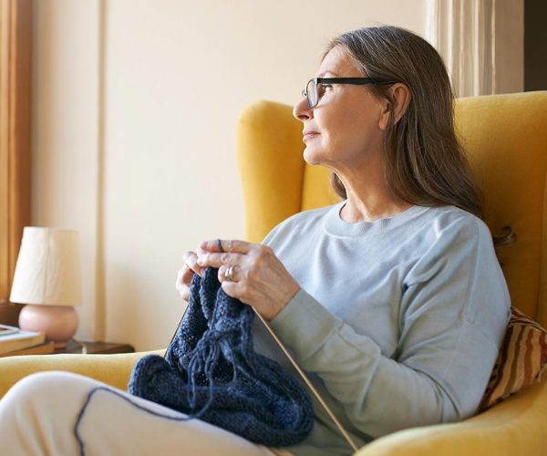 A woman, seated on a yellow armchair, is knitting with blue yarn, gazing out a window in a softly lit room with a small table lamp nearby.