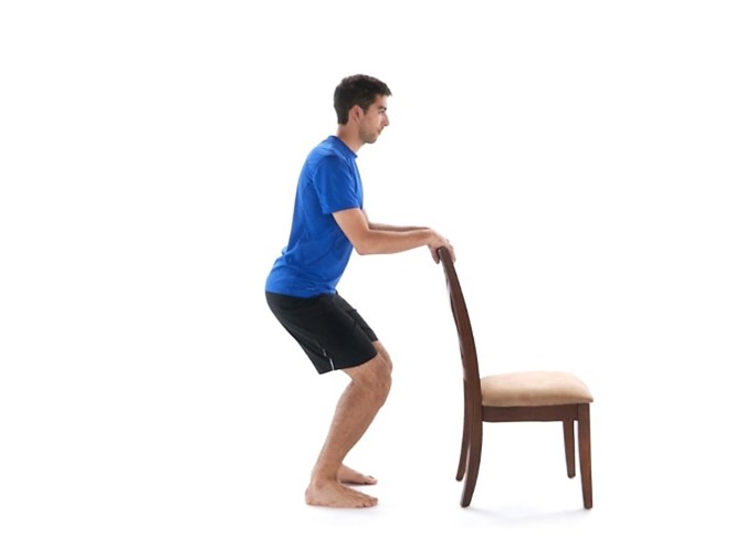 A man wearing a blue shirt and black shorts performs a squat exercise, holding onto a wooden chair for support, against a plain white background.