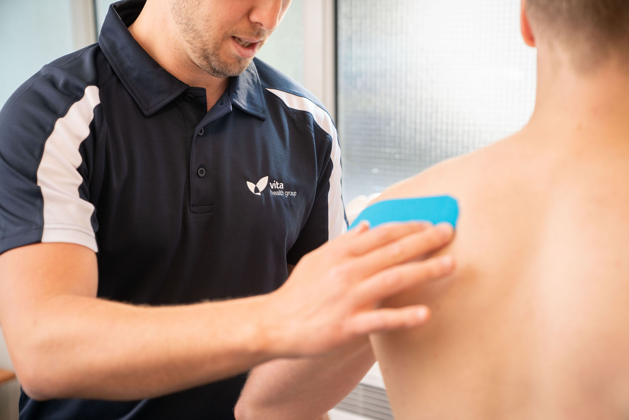 A healthcare professional applies a blue therapeutic tape to a patient's back shoulder. The professional wears a Vita Health Group polo shirt, indicating a clinical setting.
