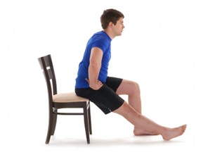 A person in a blue shirt and black shorts sits on a wooden chair, leaning forward with hands on their hips, legs extended forward in a white space.