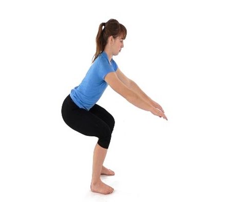 Woman performing a squat with arms extended forward in a white studio setting, wearing a blue shirt and black leggings.