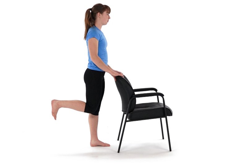 A woman stands on one leg, holding a chair for balance, performing a leg exercise. She's in a simple, unobtrusive indoor setting, wearing a blue shirt and black shorts.