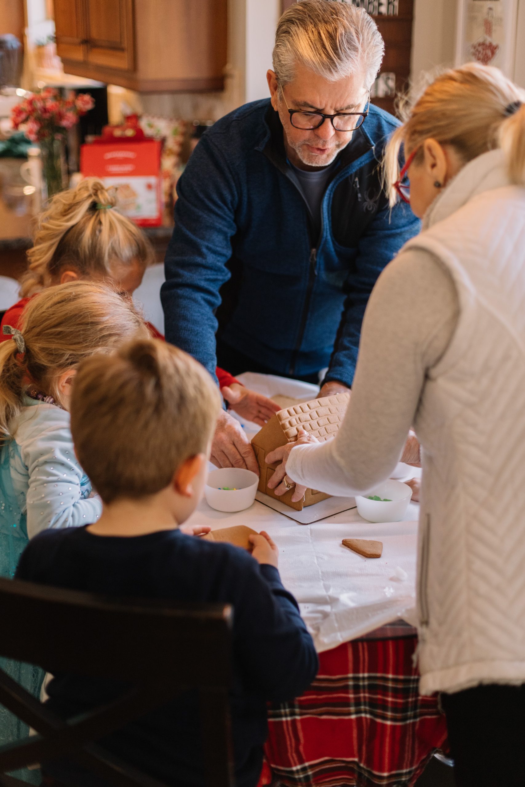 Adults and children assemble a gingerbread house on a table covered with a white cloth and bowls, in a cozy kitchen setting with wooden cabinets and festive decor.
