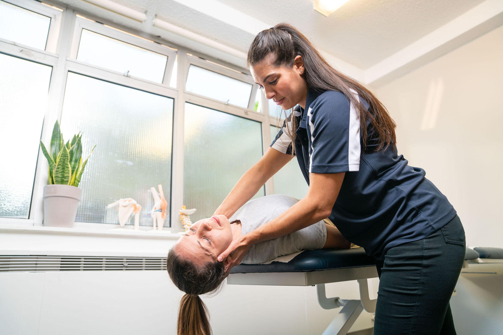 A healthcare professional assists a patient with a leg stretch on a treatment table. Sunlight streams through large windows in a bright, modern clinic setting.