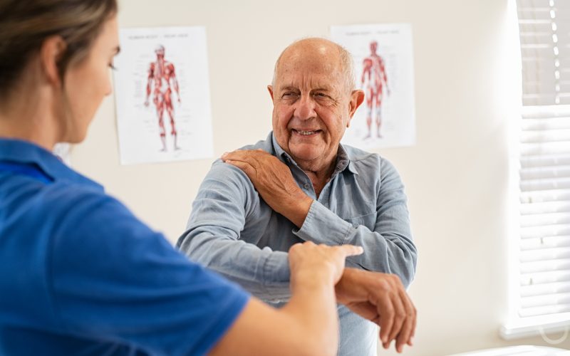 A healthcare professional guides an older man in an arm exercise. They are in a bright room with anatomy posters on the wall, enhancing a clinical atmosphere.