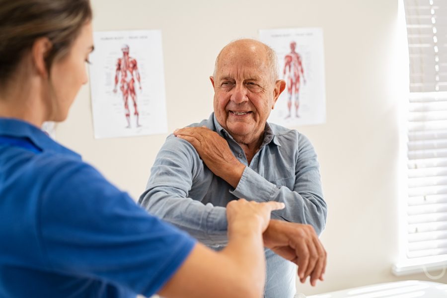 A healthcare professional guides an older man in an arm exercise. They are in a bright room with anatomy posters on the wall, enhancing a clinical atmosphere.