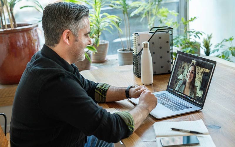 A man sits at a wooden desk engaging in a video call on a laptop. The room has large windows with plants visible in the background, creating a bright, natural atmosphere.